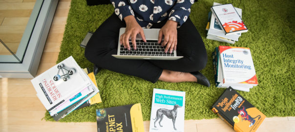 Woman types on laptop code books surround her photo by #WOCinTech Chat